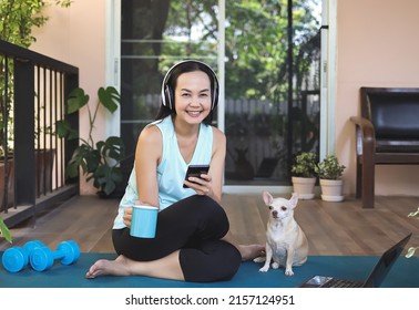 Asian woman wearing headphones, holding coffee cup, using mobile phone, sitting  on yoga mat in balcony  with computer laptop Chihuahua dog, smiling and looking at camera. Yoga training online