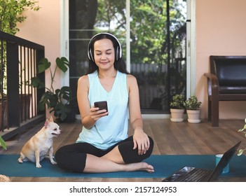 Asian woman wearing headphones,  using smartphone, sitting  on yoga mat in balcony  with computer laptop  and Chihuahua dog. Yoga exercise training online
