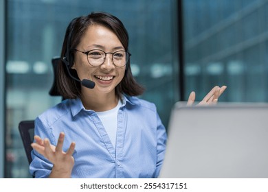 Asian woman wearing a headset is actively participating in a virtual meeting. She is gesturing with her hands and smiling, reflecting communication and engagement in a professional environment. - Powered by Shutterstock