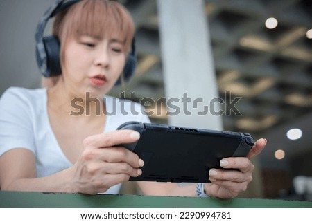 Asian woman wearing headset and playing videogame on a portable gaming console at cafe. 