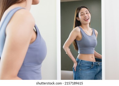 Asian woman wearing her old pant before loss weight looking reflection in the mirror and feeling joyful with her body after diet success