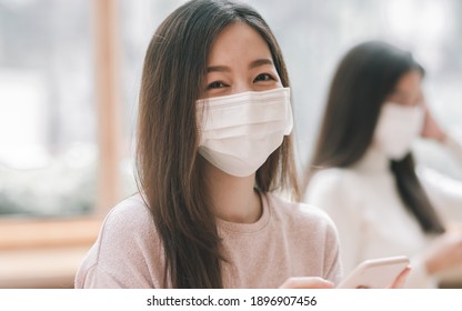 Asian Woman Wearing Mask To Protect Virus While Using Mobile Phone In Coffee Shop. New Normal And Social Distancing Concept.