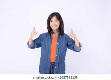 An Asian woman wearing orange t shirt and denim jean is posing with thumbs up gesture on a white background.