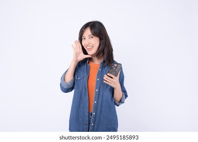 An Asian woman wearing orange t shirt and denim jean is posing with a shouting gesture while holding smartphone against a white background. - Powered by Shutterstock