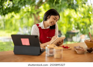 Asian woman wearing a red apron is taking notes while having a phone call in a kitchen full of fresh ingredients - Powered by Shutterstock