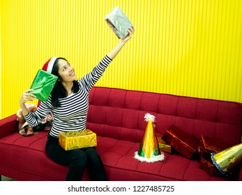 Asian woman wearing red hat feeling happy and holding gift boxes in her hands. Sitting on the red sofa in the living room. Christmas holiday party concept. - Shutterstock ID 1227485725