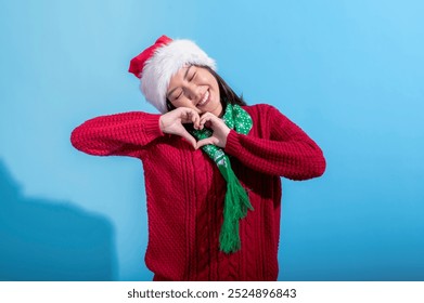 An Asian woman wearing a red sweater, green scarf with snowflake patterns, and a Santa hat, makes a heart shape with her hands in front of her chest. She smiles warmly against a light blue background - Powered by Shutterstock