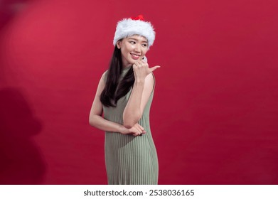 An Asian woman wearing a Santa hat and a sleeveless green ribbed dress is looking over her shoulder, gesturing with her thumb pointing backward. She smiles in front of a solid red background - Powered by Shutterstock