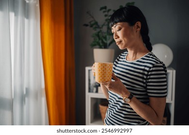 Asian woman wearing a striped shirt stands next to a window in her modern home office, holding a yellow coffee mug, looking thoughtful, surrounded by tasteful decor such as plants and shelves. - Powered by Shutterstock