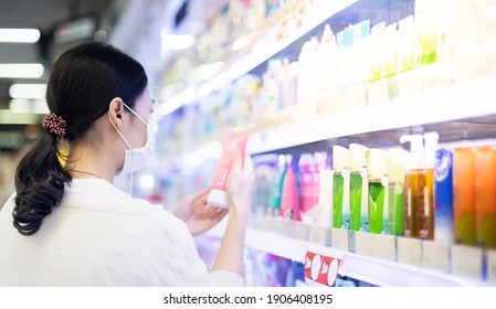 Asian Woman Wearing Surgical Mask And Hand Holding The Bottle Of Facial Foam For Shopping In The Aisle Of The Skincare Department In Supermarket, Due To The Covid-19 Pandemic Crisis.