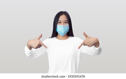 Asian Woman Wearing Surgical Mask Pointing Down Or At Her Light Clothes On White Wall Background