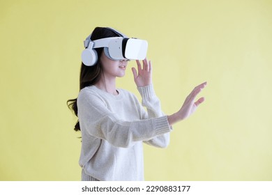 Asian woman wearing virtual reality, VR headset and trying to touch something with hand while stand over isolated yellow background - Powered by Shutterstock