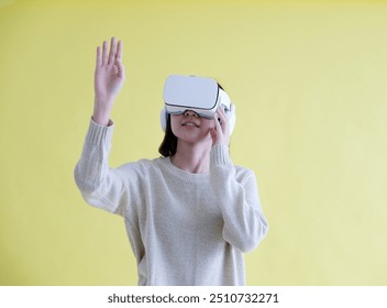 Asian woman wearing virtual reality, VR headset and trying to touch something with hand while stand over isolated yellow background - Powered by Shutterstock