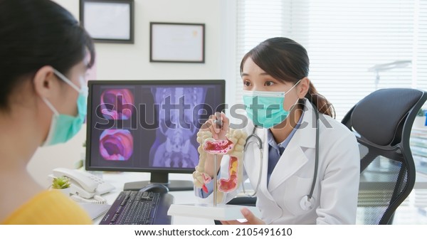 asian woman wears face mask to prevent COVID19 has\
colorectal cancer diagnosis in hospital - female doctor showing\
colonoscopy results and xray to patient on computer and explain by\
anatomical model