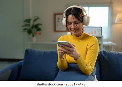 Asian woman wears headphones and selects music, watches movies, chats via social media ordering products from her smartphone and relaxing on the sofa at home, technology concept, weekend relaxation. - Powered by Shutterstock