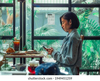 Asian Woman While Using A Smartphone, Ignoring The Food On The Table In The Restaurant. A Young Asian Woman Looking Smartphone At Her Dining Table. Side View Of Women Using The Phone.