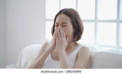 Asian Woman In White Allergy And Sneezing On Sofa In Living Room Day Time. Irritated Nose Sneeze. Authentic Lifestyle At Home.