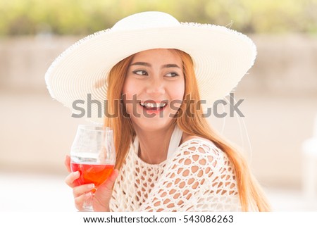 Asian woman with white large summer hat sitting on white pool bench, with cocktail near pool