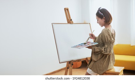 An Asian woman who loves art or a female artist is holding a paint palette and paintbrushes while happily painting on white paper at home. - Powered by Shutterstock