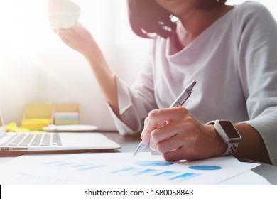 Asian Woman Work At Home Or Internet Cafe  By Holding Pen To Writing Information To Prepare Data For The Presentation, Sale Report For Meeting, Shopping Online, Chat And Hold Cup Of Coffee For Break 