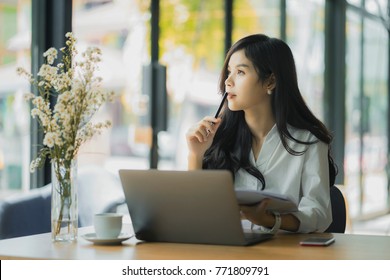 Asian Woman Working With Laptop In Coffee Shop Cafe,she Thinking And Holding Pen