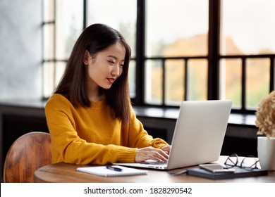 Asian Woman Working On Laptop At Home Or In Cafe. Young Lady In Bright Yellow Jumper Is Sitting At Desk Typing On Computer. Business Oriental Female In Front Of Window