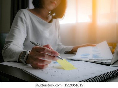Asian woman is writing on yellow paper notes and put a warning on calendar. The other hand hold the report graph and has laptop on the front. Maybe preparing data for marketing report. window light