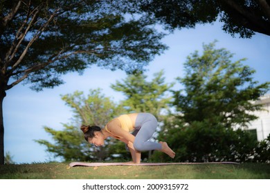 An asian woman in yellow sport bra and gray legging doing yoga in crow pose at, park under camber of big tree. Yoga practicing in nature, greenery, and freshness.
