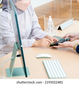 Asian Woman's Hands, Entering Pin Code On A Pin Pad. Woman Bank Teller Holding A Credit Card. A Sanitizing Gel And Acrylic Divider As Part Of Safety Protection For Customers. Covid-19 Health Protocol.