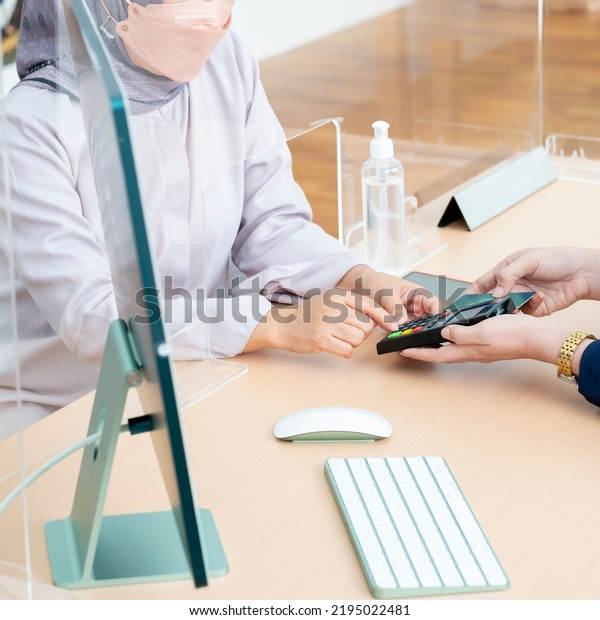 Asian woman\'s hands, entering pin code on a pin\
pad. Woman Bank teller holding a credit card. A sanitizing gel and\
acrylic divider as part of safety protection for customers.\
Covid-19 health protocol.