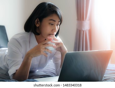 Asian woman's tan skin and short black hair with wearing white shirt on grey bed and hold white coffee cup in hand. See the screen laptop with serious on face may check report or just use social media - Shutterstock ID 1483340318