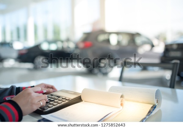 Asian women Car salesman calculating a price\
at the dealership office. pressing calculator for business finance\
on car showroom blurry background.\
