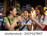 Asian women friend eating street food while traveling in city at night. Attractive girls tourist feeling happy and relax, walking on street enjoy spend free leisure time for holiday vacation together.