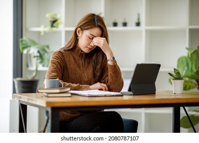 Asian women sitting in a home office With stress and eye strain.Tired businesswoman holding eyeglasses and massaging nose bridge. There are tablets, laptops, and coffee. - Shutterstock ID 2080787107