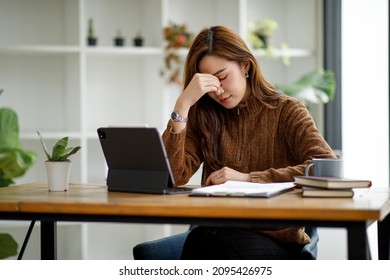 Asian women sitting in a home office With stress and eye strain.Tired businesswoman holding eyeglasses and massaging nose bridge. There are tablets, laptops, and coffee. - Shutterstock ID 2095426975