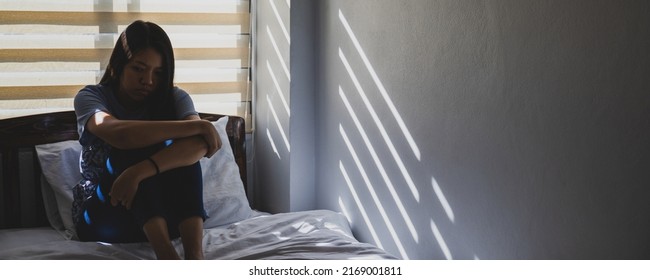 Asian women are sitting hugging their knees in bed. Feeling sad, disappointed in love In the dark bedroom and sunlight from the window through the blinds.