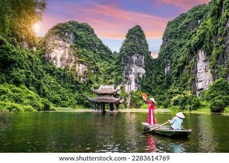 Asian women wearing red traditional Vietnamese cultural clothing on a boat floating in the river flowing through the mountains at Trang An, Vietnam.