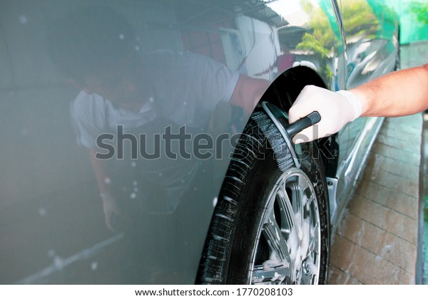 Asian worker\
in car care garage cleaning a dirty vehicle wheels by using brush\
and liquid soap foam close up with copyspace. Skilled Asian labor\
working in car care service\
concept.