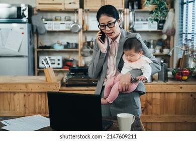 asian working lady standing and looking down at computer is discussing business on phone and caring her kid. chinese career woman carrying baby is answering call and viewing data on notebook at home.