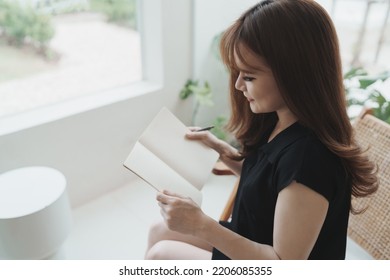 Asian Working Woman Wearing Black Shirt And Writing Journal On Small Notebook On The Table At Indoor Cafe. Woman Notes And Drinking Coffee At Cafe. Working From Anywhere Concept.