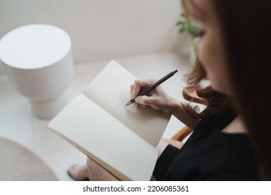 Asian Working Woman Wearing Black Shirt And Writing Journal On Small Notebook On The Table At Indoor Cafe. Woman Notes And Drinking Coffee At Cafe. Working From Anywhere Concept.