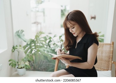 Asian Working Woman Wearing Black Shirt And Writing Journal On Small Notebook On The Table At Indoor Cafe. Woman Notes And Drinking Coffee At Cafe. Working From Anywhere Concept.