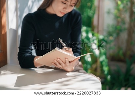 Asian working woman wearing black shirt and writing journal on small notebook on the table at indoor cafe. Woman notes and drinking coffee at cafe. Working from anywhere concept.