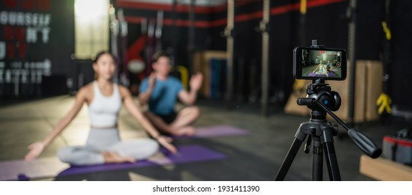 Asian Yoga Woman Teacher Teaching Caucasian Man In Gym. The Girl Broadcast Her Class Live Online And Recording By Smartphone To Quarantine Student At Home When Fitness Close During COVID 19 Pandemic.