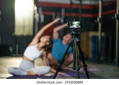 Asian Yoga Woman Teacher Teaching Caucasian Man In Gym. The Girl Broadcast Her Class Live Online And Recording By Smartphone To Quarantine Student At Home When Fitness Close During COVID 19 Pandemic.