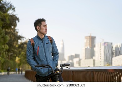 Asian Young Adult Man Sitting On His Bicycle Lost In Thought In City Park