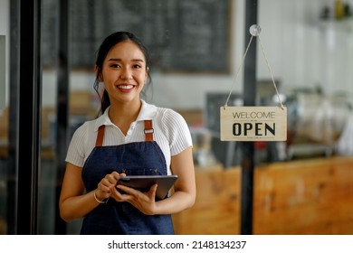 Asian Young Barista Woman In Apron Holding Tablet And Standing In Front Of The Door Of Cafe With Open Sign Board. Small Business Owner Startup Concept.