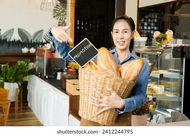 Asian young barista worker female holding Promotion sign board and bread in basket in front of bakery cabinet taking pictures to promote in social media channel, happy small business owner lifestyle - Powered by Shutterstock