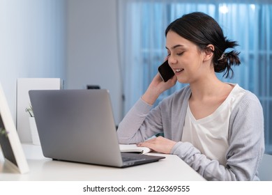 Asian Young Beautiful Businesswoman Working At Home During Night Time. Attractive Freelancer Girl Sit On Study Table, Use Mobile Phone Call For Late Work Meeting And Write Note In Living Room In House