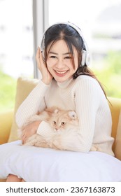 Asian young beautiful female teenager girl owner in turtleneck sweater wearing headphones listening to music sitting smiling on cozy sofa holding cute little orange long hair domestic cat on pillow. - Shutterstock ID 2367163873